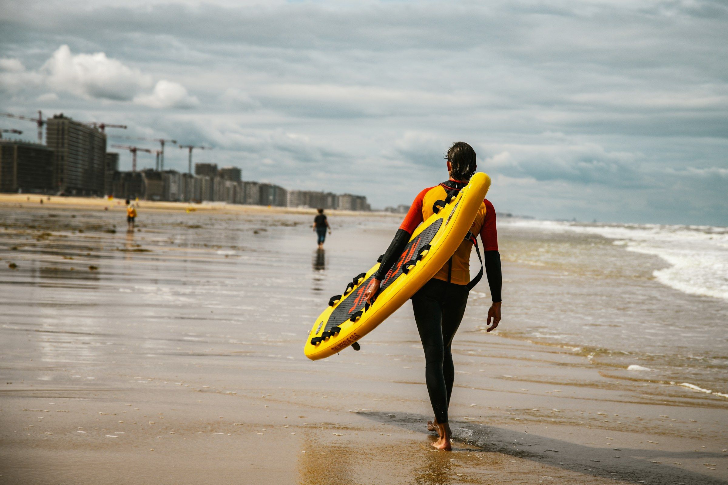 Une sauveteuse en combinaison marche au bord de l’eau, planche de sauvetage jaune sous le bras