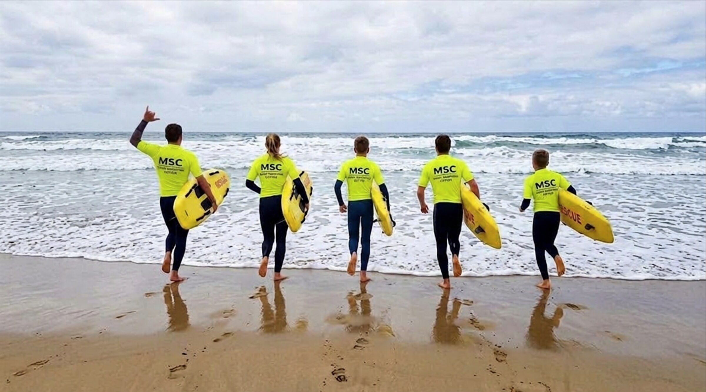 Cinq sauveteurs en lycra jaune « MSC » courent vers l'océan, planche de sauvetage sous le bras